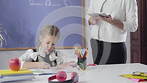 Unrecognisable female caucasian tutor teaching cute girl at home. Intelligent child studying with private teacher