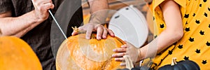 Unrecognisable young girl sitting on kitchen table, helping her father to carve large pumpkin. Halloween family banner.