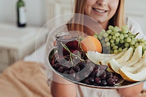 Unrecognisable Young Woman Holding Tray of Fruit in Hotel Room, Bottle of Wine Stands on the Background