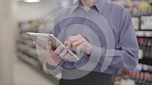 Unrecognizable adult Caucasian man using tablet at the background of blurred shelves with alcohol in supermarket. Male