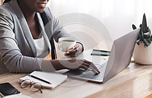 Unrecognizable afro businesslady working on laptop and drinking coffee at workplace