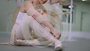 Unrecognizable ballet dancer tying laces on pointe shoes sitting on floor in dance studio. Blurred people at barre at