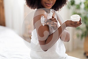 Unrecognizable black lady applying body cream on shoulder after shower in morning at home