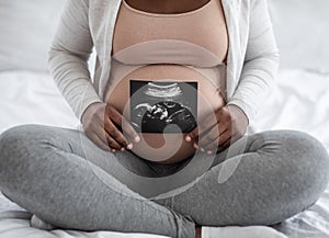 Unrecognizable black pregnant lady demonstrating her baby sonography photo, sitting on bed