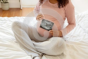 Unrecognizable black pregnant woman showing baby sonography photo while sitting on bed