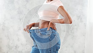 Unrecognizable black woman in oversized jeans showing results of her weight loss diet or liposuction, light background