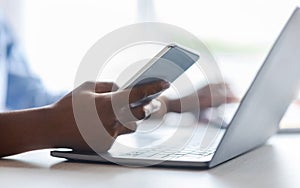 Unrecognizable black woman using smartphone and working on laptop in office, closeup