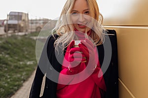 Unrecognizable blond ladylike woman warming with hot cup of coffee, looking at camera on cold balcony. Take away coffee