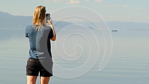 Unrecognizable blonde woman in blue shirt and black short shorts using her phone taking pictures of lake or sea. rowboat