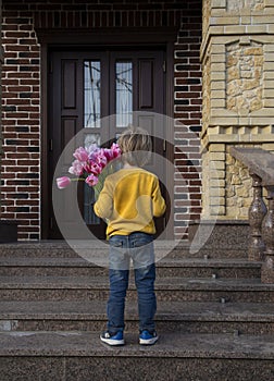 Unrecognizable boy of 5-6 years old stands with his back with a bouquet of tulips in front of the door of house