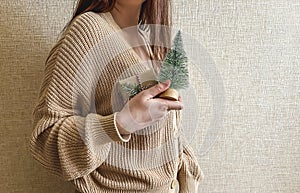 An unrecognizable brunette girl in a beige sweater holds a Christmas gift in eco packaging and a green Christmas tree in her hands