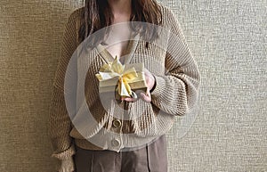 An unrecognizable brunette girl in a beige sweater holds a gift with a ribbon in her hands