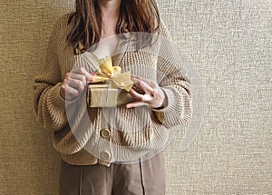 An unrecognizable brunette girl in a beige sweater holds a gift with a ribbon in her hands.
