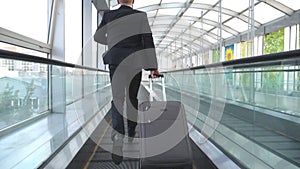 Unrecognizable businessman with suitcase on rolls runs on escalator in hall of terminal. Young man is late for flight