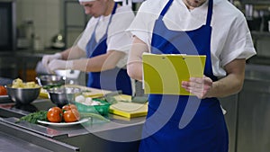 Unrecognizable chef in apron writing new recipe standing in kitchen with blurred cook working at background. Young