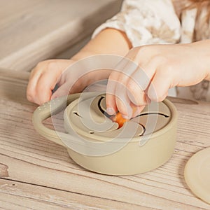 Unrecognizable child taking piece of carrot out from pastel gray silicone snack cup near lid at wooden table. Tableware.