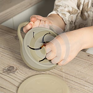 Unrecognizable child taking piece of snack from pastel gray silicone snack cup near lid at wooden table. Baby tableware.