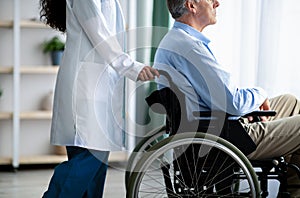 Unrecognizable elderly male patient in wheelchair and his female caregiver at retirement home, cropped view