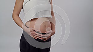 Unrecognizable expectant mother gently caressing her abdomen with baby on standing white background. Pregnant woman