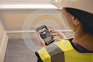 Unrecognizable female civil engineer calculating with a calculator in a construction site, wears yellow high visibility vest