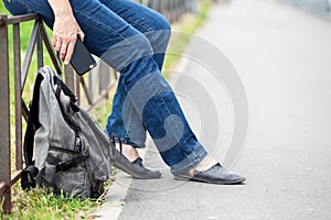 Unrecognizable female sitting on steel fence of pedestrian pathway with mobile phone in hands, urban backpack standing on earth