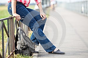 Unrecognizable female sitting on steel fence of pedestrian pathway with urban backpack on earth