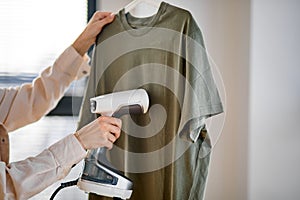 Unrecognizable female using electric steamer at home, ironing clothes on hanger