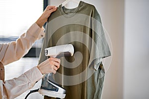 Unrecognizable female using electric steamer at home, ironing clothes on hanger photo
