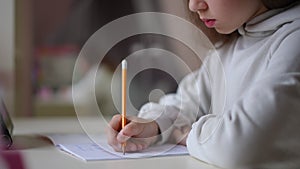 An unrecognizable girl sits at a table, on which the tablet sits, and writes with a pencil in a exercise book. A child