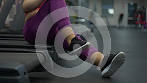 Unrecognizable heavyset Caucasian woman sitting on treadmill resting after training. Overweight plus-size sportswoman