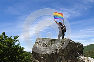 An unrecognizable hiker with a backpack, perched on a rock at the top of a mountain holding a rainbow flag fluttering in the wind