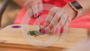 Unrecognizable lady cutting greens on wooden board outdoors. Woman's hands with bright manicure cutting verdure with