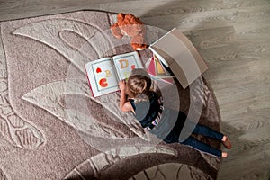 Unrecognizable little girl in a dark blue dress reading book lying on the floor near teddy bear. Child reads story for toy. Top v