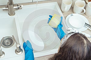 Unrecognizable little preteen girl wearing big blue rubber gloves, holding soapy sponge, washing white plate in kitchen.
