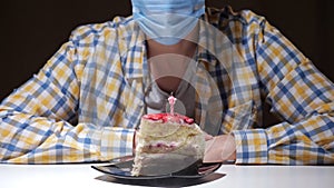 unrecognizable man in blue disposable facial mask blows out fire on candle of birthday cake put on plate sitting alone