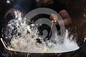 Unrecognizable man cooking in fatiscent big pan or wok in a small street food stall. White smoke coming out from the pan, hand and