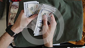 Unrecognizable man hands with pile of money, US dollars notes - counting under the suitcase with clothes. Counting money