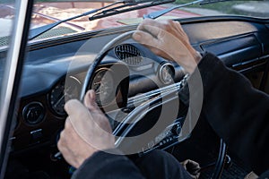 Unrecognizable man holding hands on steering wheel and driving retro car through country road at sunny autumn day. Guy