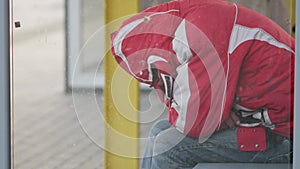 An unrecognizable man in a red jacket sits at a public transport stop. No face