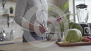 Unrecognizable man in the shirt cooking soup checking recipe on the tablet in the kitchen at home. Concept of healthy