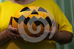 An unrecognizable man in a yellow t-shirt holds an orange Jack pumpkin with a gruesome painted face for Halloween. Angry smiling p