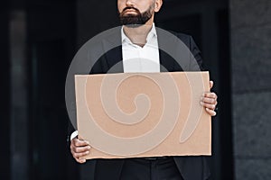 Unrecognizable middle-aged businessman in suit holding blank cardboard sign standing outdoors, empty space