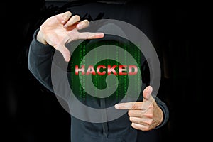 Unrecognizable person in a black hoodie shows a green number matrix displaying the text Hacked