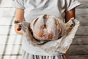 unrecognizable person in neutral light clothing holds round  freshly baked brown grain bread in hands. Products home bakery 