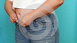 Unrecognizable plump woman wear beige underwear, white T-shirt, pull on jeans, trying to button up on blue background.