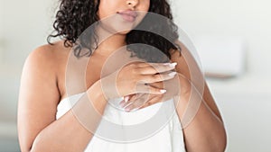 Unrecognizable plus size lady in white towel applying moisturising lotion or nourishing cream on hands, home interior