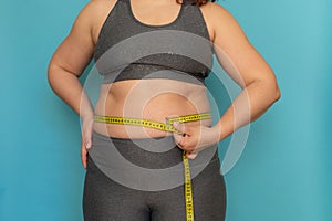 Unrecognizable plus-size woman in grey sports bra and leggings, showing excess naked belly, measuring waist with tape.