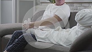 Unrecognizable positive mature man sitting on the sofa and massaging wife`s feet. Husband taking care of his senior wife