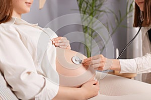 Unrecognizable pregnant female with bare belly and stethoscope doctor listening to baby heartbeat woman wearing white shirt