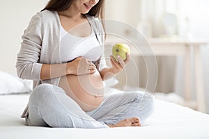 Unrecognizable pregnant lady sitting on bed, holding green apple